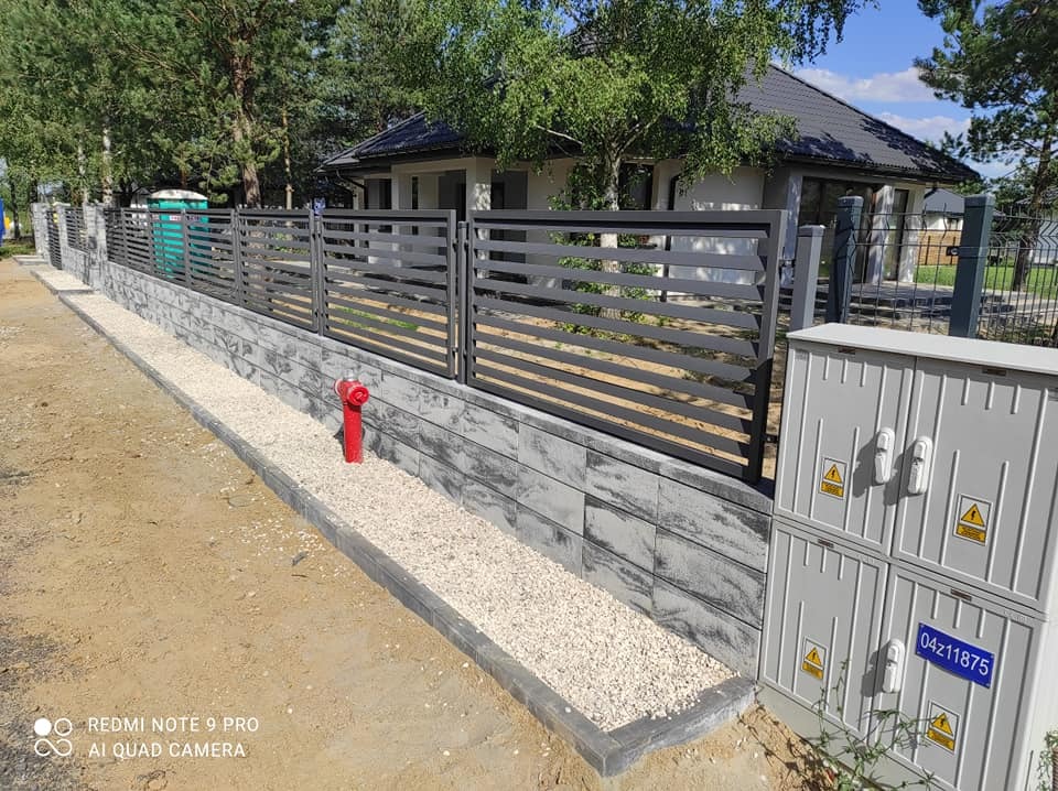 Nowoczesne ogrodzenie panelowe w kolorze antracytowym, osadzone na murku z szarej kostki łupanej, z podsypką z białego grysu, na tle domu jednorodzinnego.