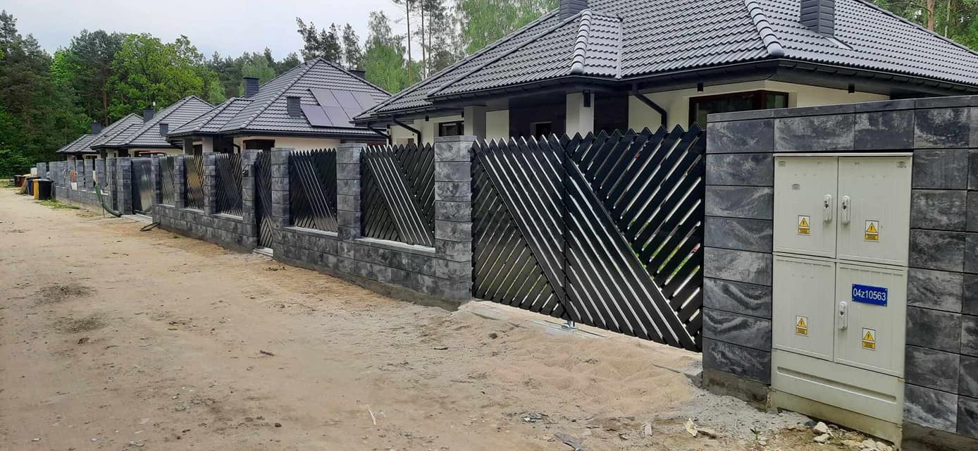 Nowoczesne ogrodzenie posesji z szarej kostki łupanej i czarnych, ukośnych paneli drewnianych, widok z zewnątrz na tle domów z ciemnymi dachami i zieleni drzew.