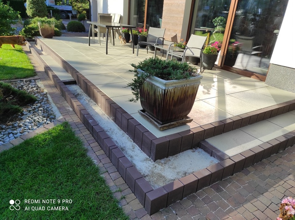 Jasne, kwadratowe płyty tarasowe z ciemnobrązowym obramowaniem z kostki, prowadzące do domu z dużymi oknami; donica z zieloną rośliną na pierwszym planie, trawnik i ogród w tle.