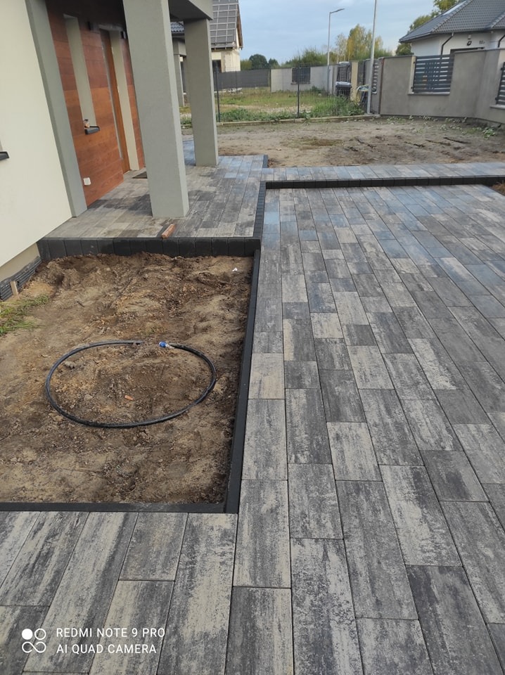 Częściowo ułożona nawierzchnia z szarej kostki z czarną obwódką, widoczny fragment domu i rabata z czarnym wężem ogrodowym.
