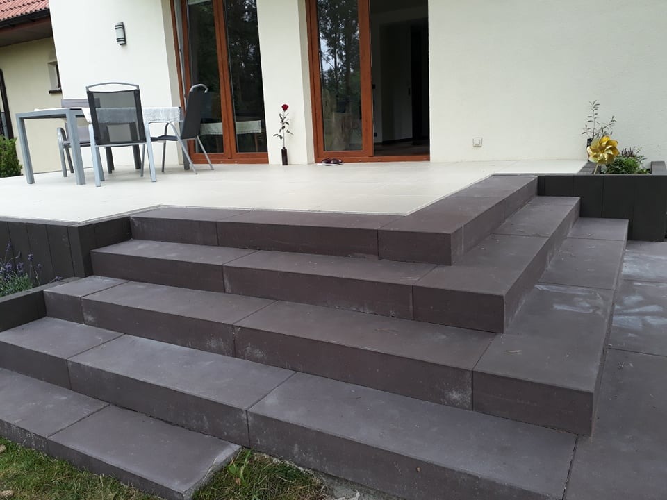 Schody z ciemnoszarej kostki prowadzące na taras domu jednorodzinnego, z elementami dekoracyjnymi w postaci roślin w donicach i metalowej wiatraczka.