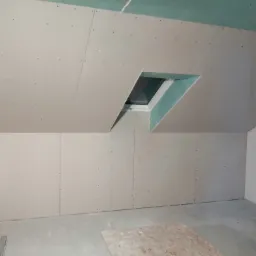 Poddasze w trakcie wykańczania: ściany i skośny sufit pokryte płytami gipsowo-kartonowymi, widoczne wkręty, okno dachowe w obudowie z płyt GK, na podłodze płyta OSB.