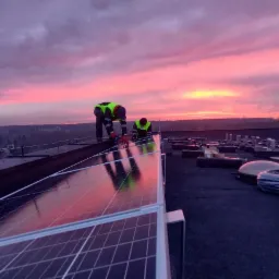 Montaż paneli słonecznych na dachu budynku podczas zachodu słońca, widoczni dwaj pracownicy w kamizelkach odblaskowych.