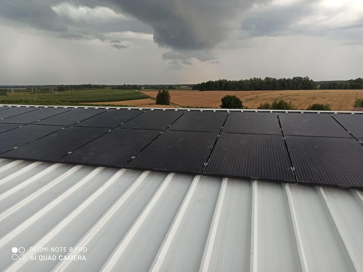 Panele słoneczne na blaszanym dachu w pochmurny dzień, widok na pole uprawne w tle.