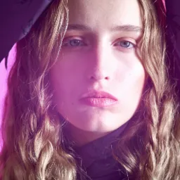 Portret młodej kobiety z blond włosami w warkoczach, oświetlonej fioletowym światłem, z ciemnym materiałem z wzorem drapowanym nad głową.