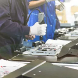 Pracownicy w niebieskich uniformach i białych rękawiczkach przy linii montażowej, używający wkrętarek akumulatorowych do składania elementów elektronicznych na jasnych stołach roboczych.