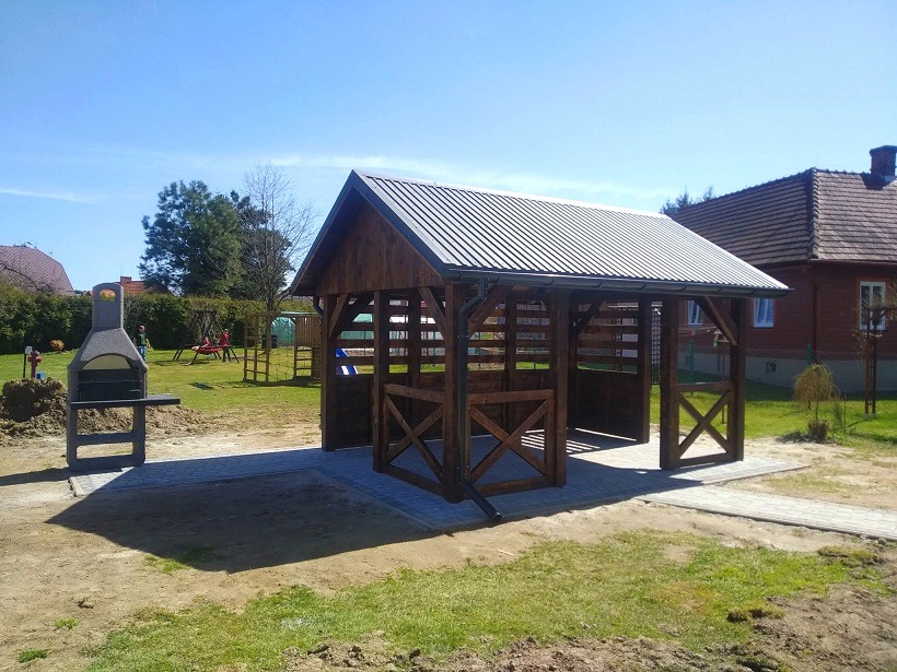 Drewniana altana z metalowym dachem, widoczna z boku na trawniku, obok stoi grill, w tle dom i drzewa.