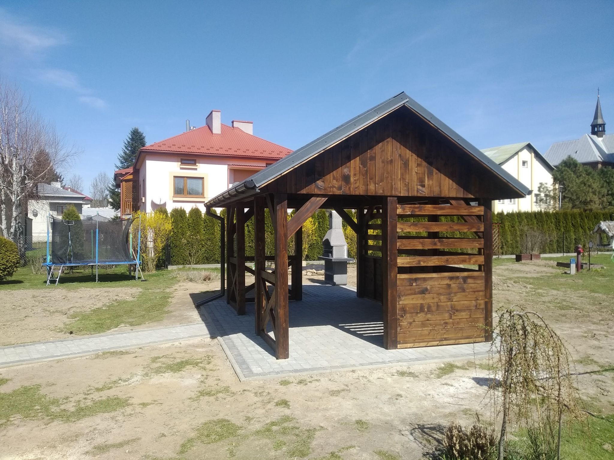 Drewniana konstrukcja z dachem na kostce brukowej, z grillem w tle, otoczona zielenią i zabudową mieszkalną w słoneczny dzień.