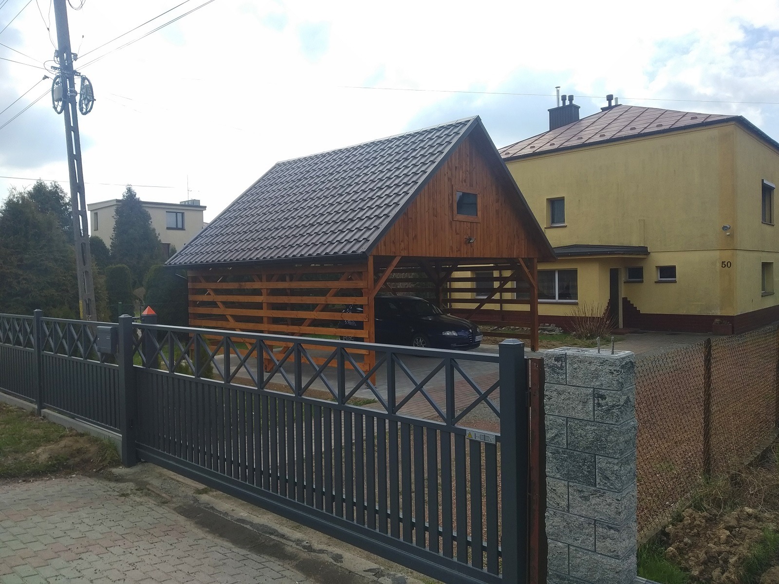 Drewniana wiata garażowa z brązowym dachem kryjąca samochód, widoczna zza szarego metalowego ogrodzenia z kamiennymi słupkami