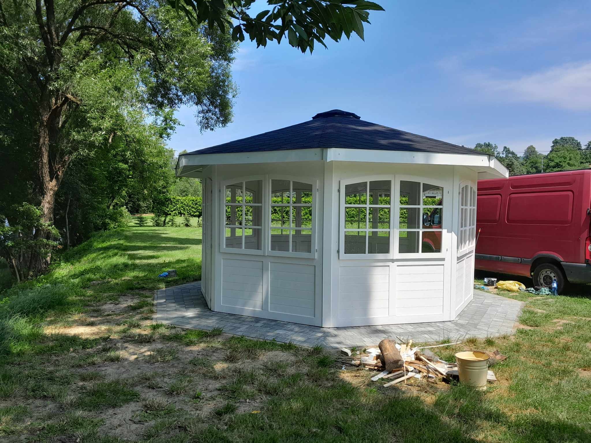 Biała, ośmiokątna konstrukcja z oknami i czarnym dachem, stojąca na szarej kostce brukowej w ogrodzie, obok leżą kawałki drewna i stoi wiadro, w tle czerwony samochód dostawczy.