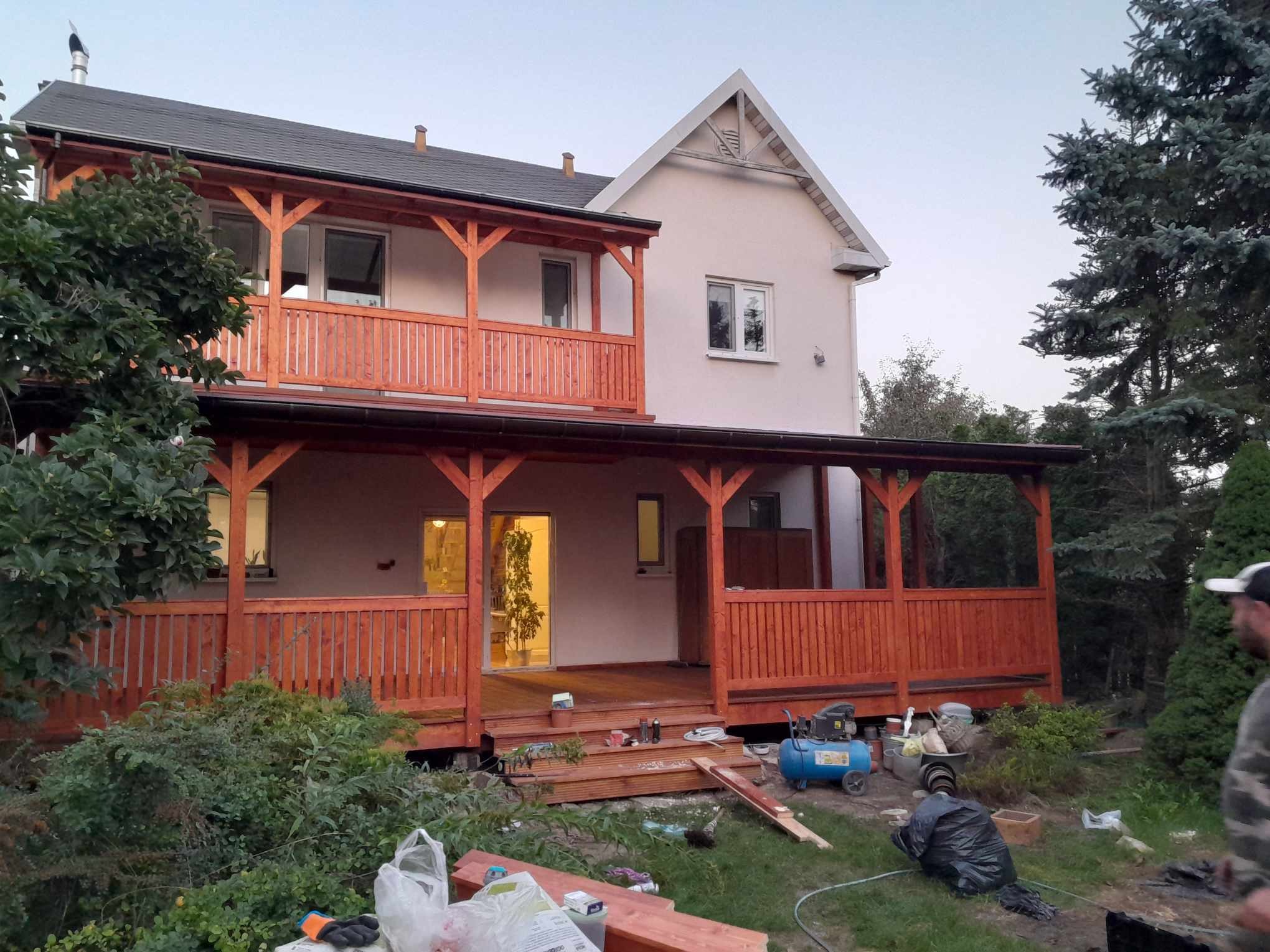 Dwukondygnacyjny dom z drewnianym tarasem i balkonem w trakcie budowy, widoczne narzędzia i materiały na trawniku.