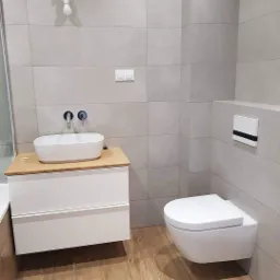 Nowoczesna łazienka z umywalką nablatową na szafce z drewnianym blatem, podwieszaną toaletą i szarymi płytkami.