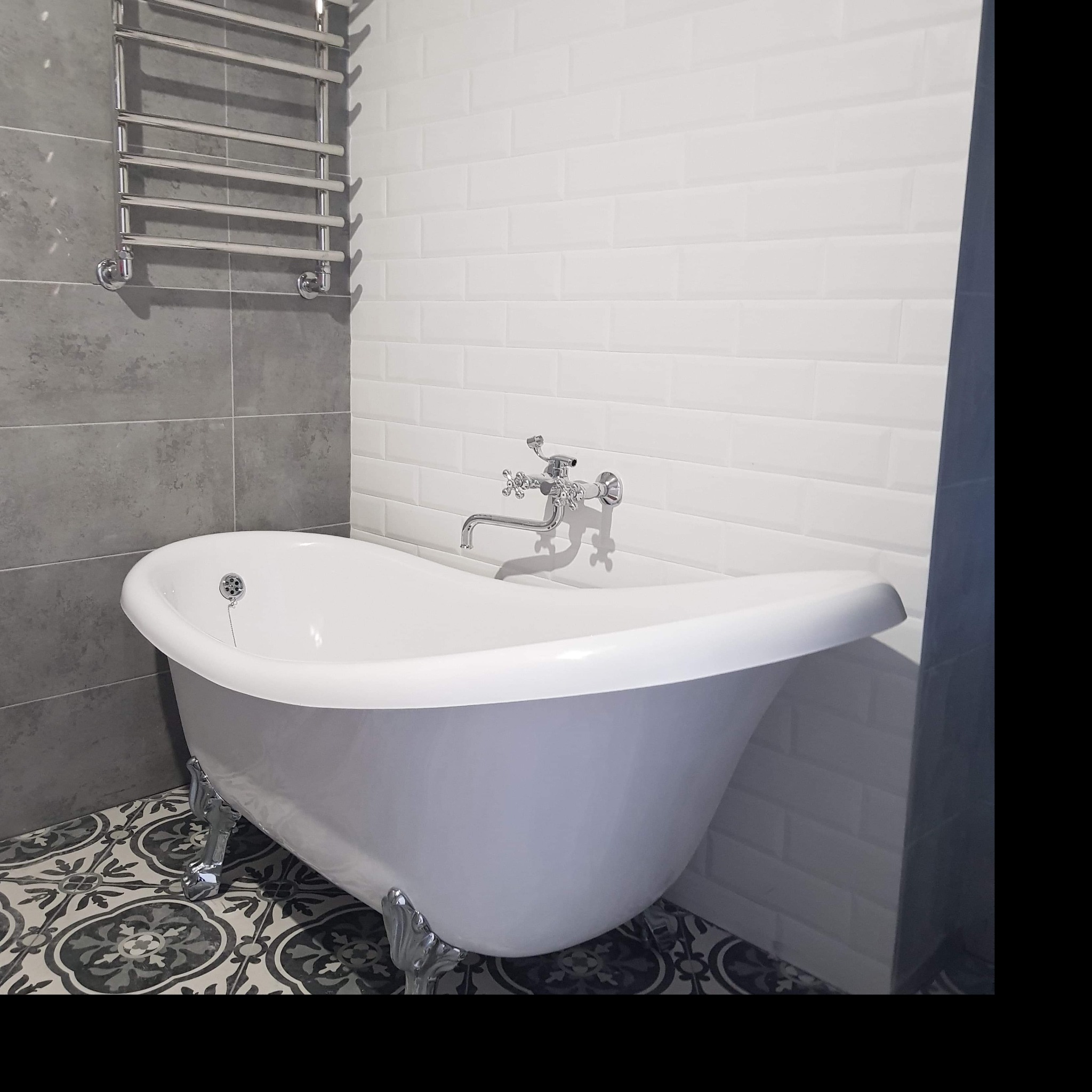 Wolnostojąca wanna na dekoracyjnych płytkach podłogowych, z chromowaną baterią i grzejnikiem drabinkowym na ścianie wyłożonej szarymi i białymi płytkami typu metro.