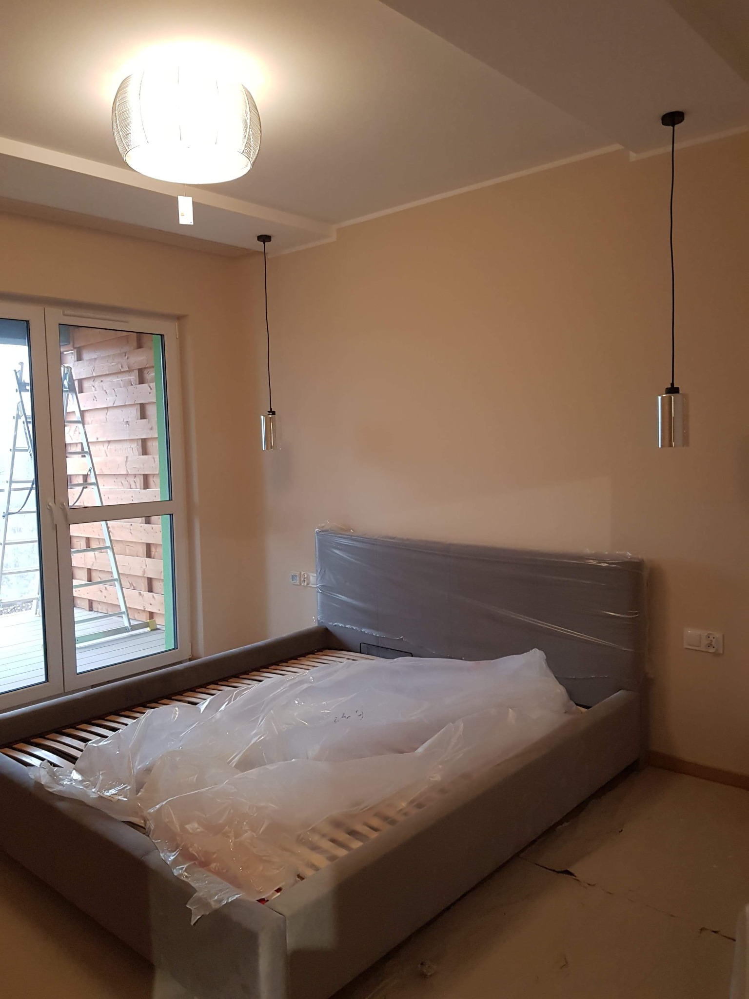 Pokój po malowaniu ścian na beżowo, z nowym szarym łóżkiem w folii, wiszącymi lampami i drabiną opartą o okno.