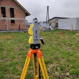 Żółto-szary tachimetr marki GeoMax ZOOM40 na pomarańczowym statywie, ustawiony na trawiastym terenie budowy z widocznym budynkiem w stanie surowym otwartym z czerwonej cegły w tle.