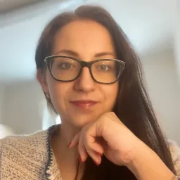 Portret kobiety w okularach, z dłonią pod brodą, ubrana w tweedowy żakiet, z delikatną bransoletką na nadgarstku.