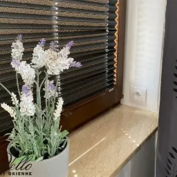 Brązowe plisy okienne zamontowane w oknie, z dekoracyjnym bukietem kwiatów w doniczce na parapecie i zasłoną z wycinanymi wzorami obok.