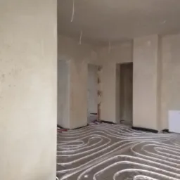 Surowe, jasne wnętrze z instalacją ogrzewania podłogowego przed wylaniem posadzki; widoczne przewody wystające z sufitu i świeżo nałożone, surowe wykończenie ścian.
