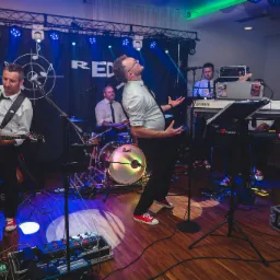 Zespół muzyczny RED na scenie: gitarzysta z białą gitarą Ibanez, wokalista w okularach z mikroportem w uniesieniu, perkusista za perkusją Pearl z podświetlanym bębnem, klawiszowcy za klawiszami...