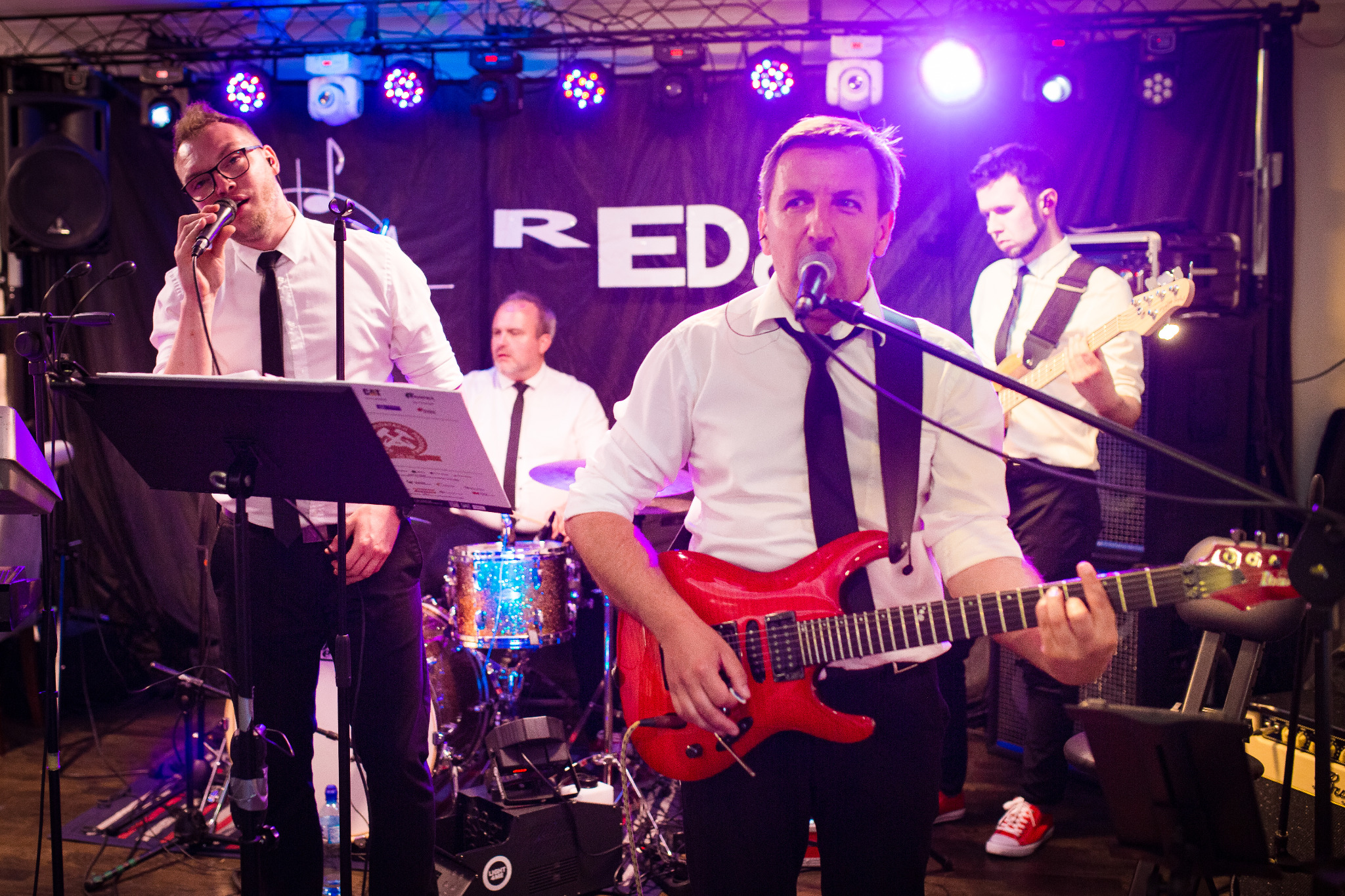 Zespół muzyczny w białych koszulach i czarnych krawatach występuje na scenie, wokalista gra na czerwonej gitarze, w tle perkusja i logo zespołu na czarnym tle, oświetlenie sceniczne w kolorze...