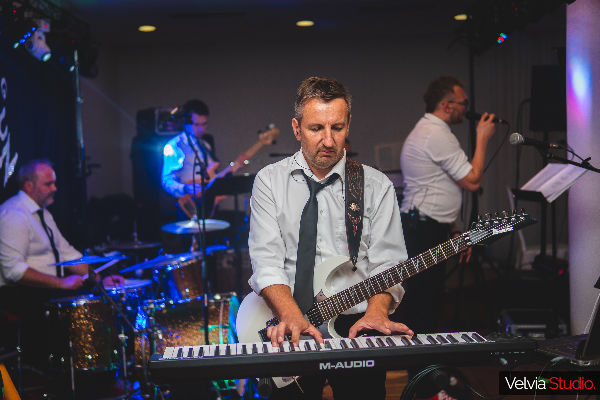 Muzyk grający na keyboardzie M-Audio z gitarą Ibanez na szyi, w tle perkusja, basista i wokalista podczas występu na scenie z oświetleniem.