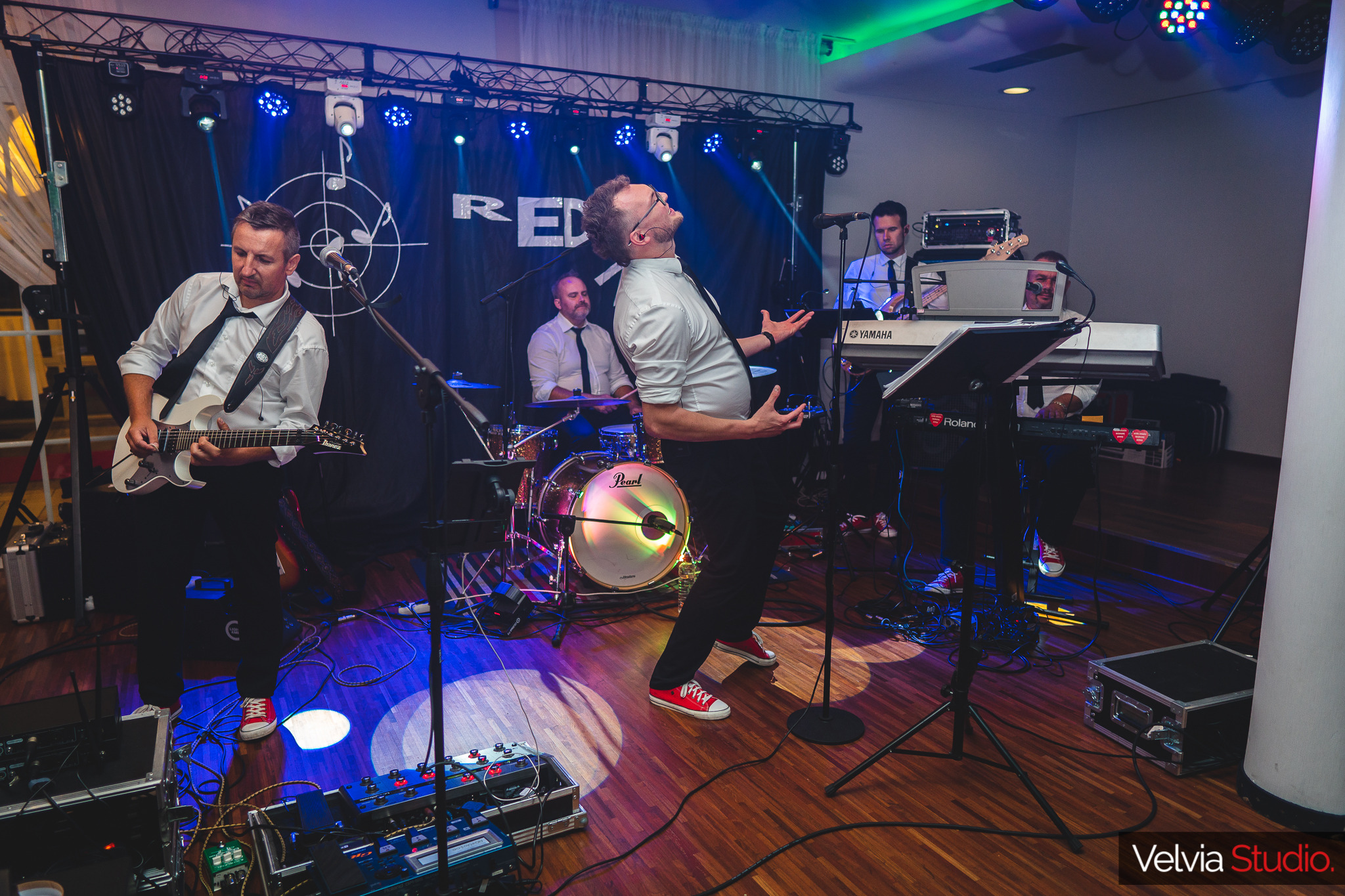 Zespół muzyczny RED na scenie: gitarzysta z białą gitarą Ibanez, wokalista w okularach z mikroportem w uniesieniu, perkusista za perkusją Pearl z podświetlanym bębnem, klawiszowcy za klawiszami...
