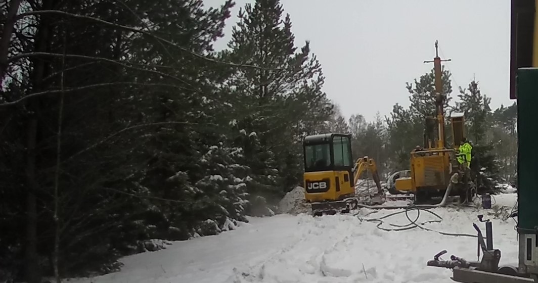 Wiercenie otworów pod pompy ciepła w zimowych warunkach; żółty mini-koparka JCB i wiertnica na tle zasypanego śniegiem lasu iglastego, pracownik w jaskrawej kurtce przy maszynie.