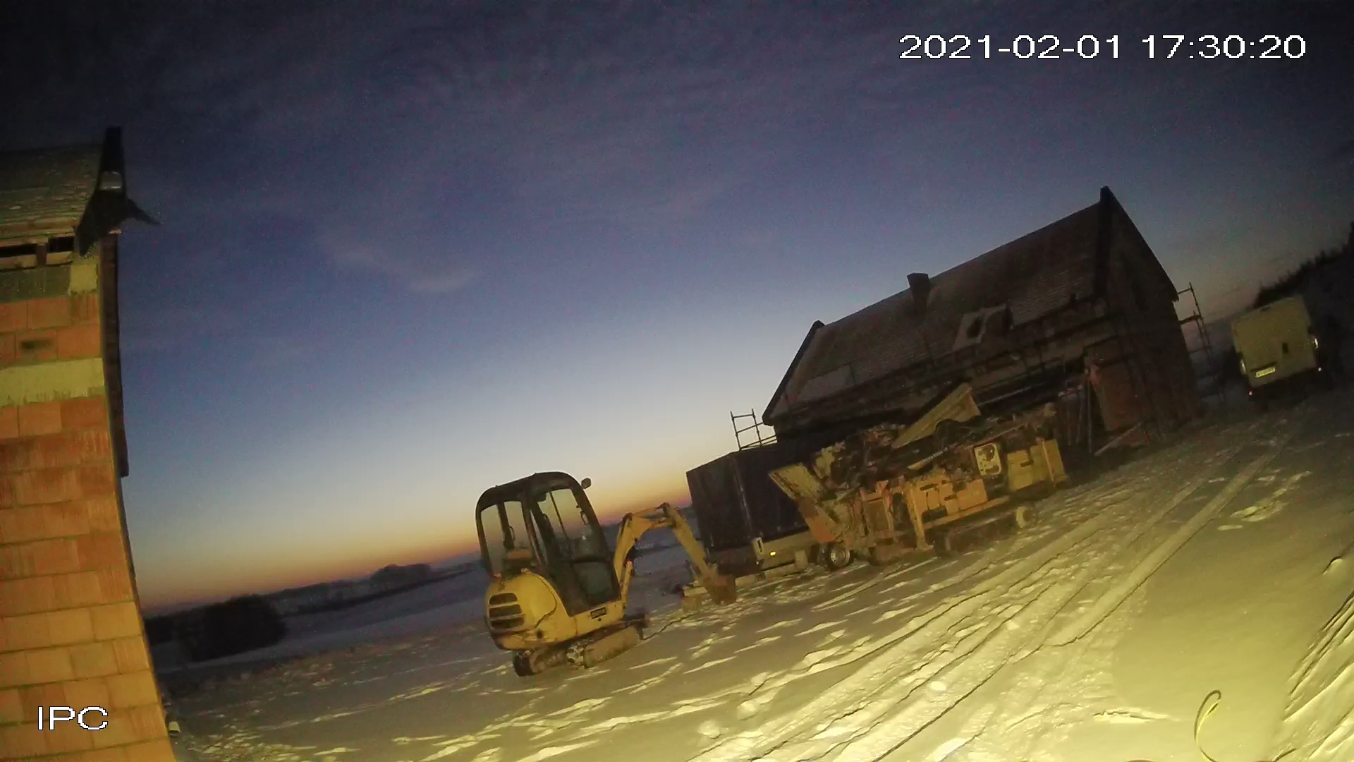 Nocne ujęcie małej koparki na śniegu przed budynkiem, zarejestrowane kamerą przemysłową z datą i godziną w rogu.