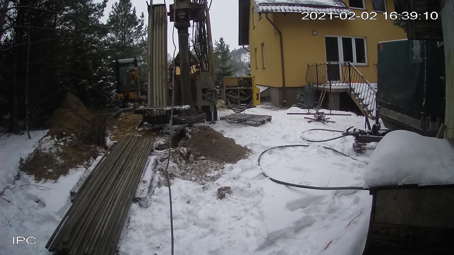 Wiercenie otworów pod instalację, widoczna wiertnica na tle zasypanego śniegiem terenu i żółtego budynku, rury wiertnicze ułożone na śniegu.