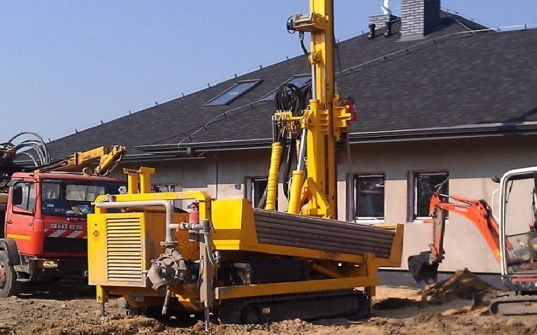 Żółta wiertnica na gąsienicach podczas prac ziemnych przy budynku z ciemnym dachem i oknami, widoczny również czerwony samochód ciężarowy i mała koparka.