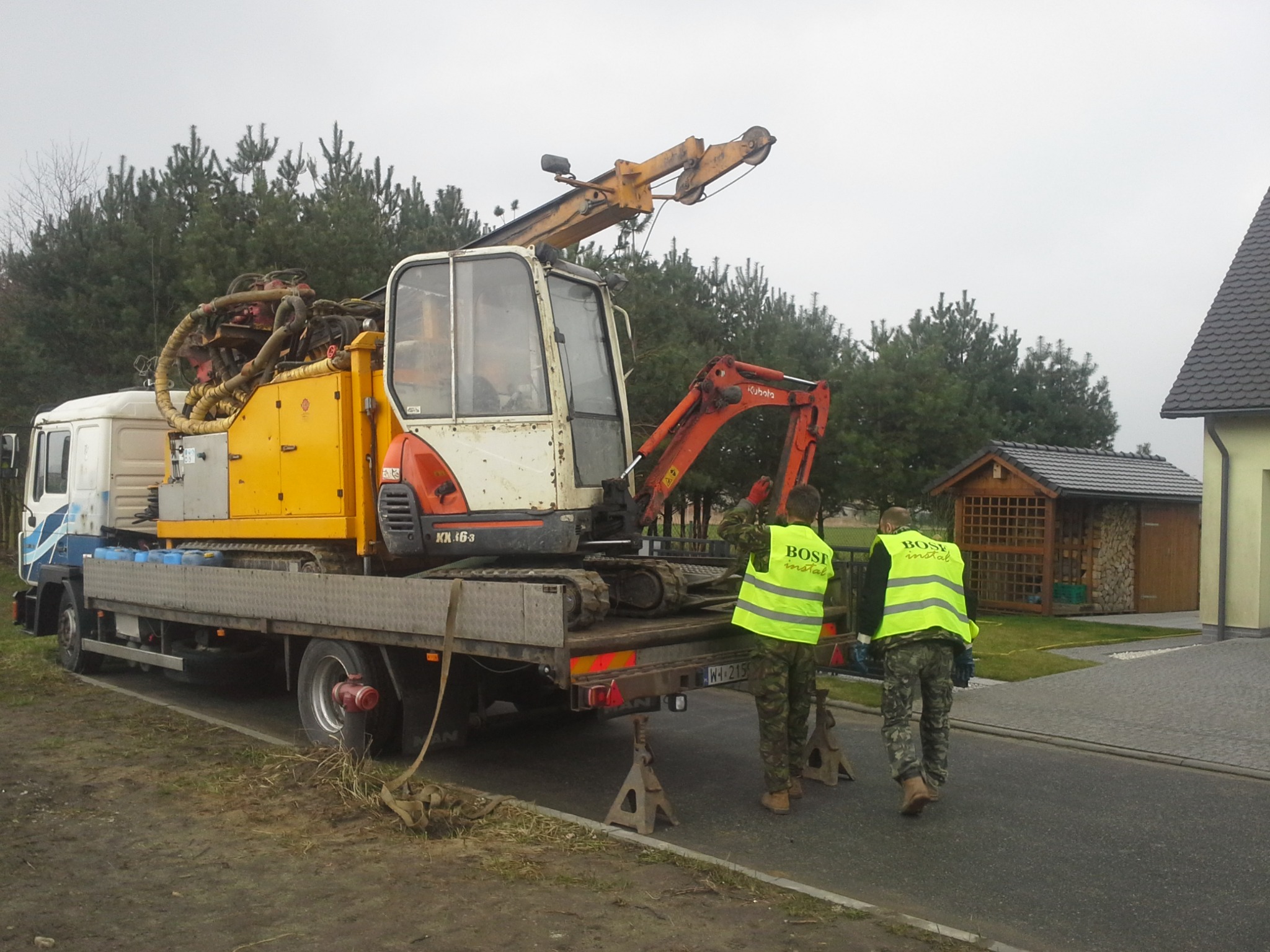 Transport wiertnicy na podwoziu gąsienicowym, załadowanej na lawetę ciężarówki, zabezpieczonej podporami, z dwoma pracownikami w kamizelkach odblaskowych na tle domów i drzew