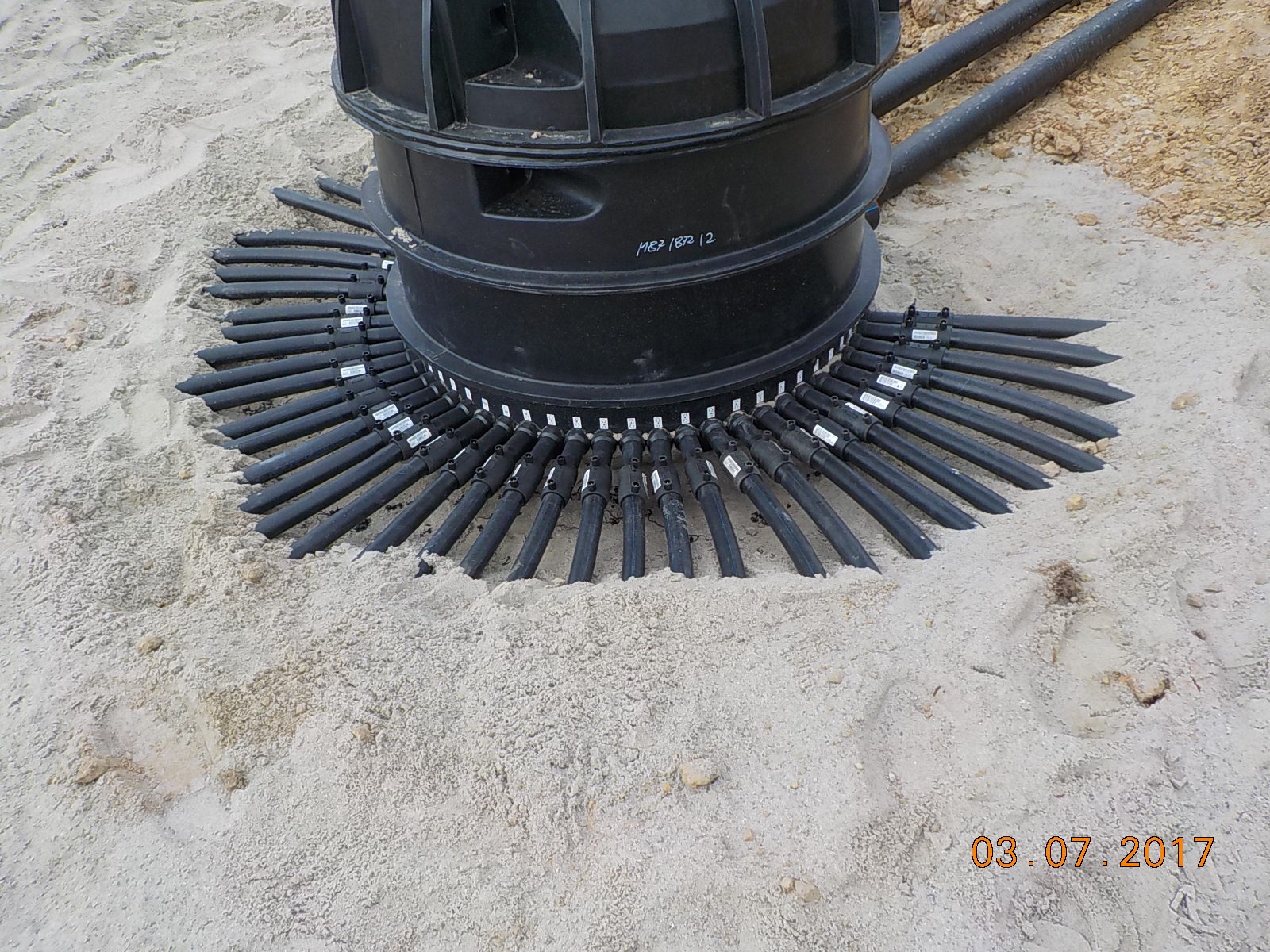 Czarny kolektor gruntowy z rurami ułożonymi promieniście w piasku, widoczny numer seryjny na obudowie.