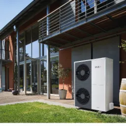 Biała pompa ciepła Vaillant aroTHERM plus umieszczona na trawniku przed nowoczesnym domem z dużymi oknami i balkonami.