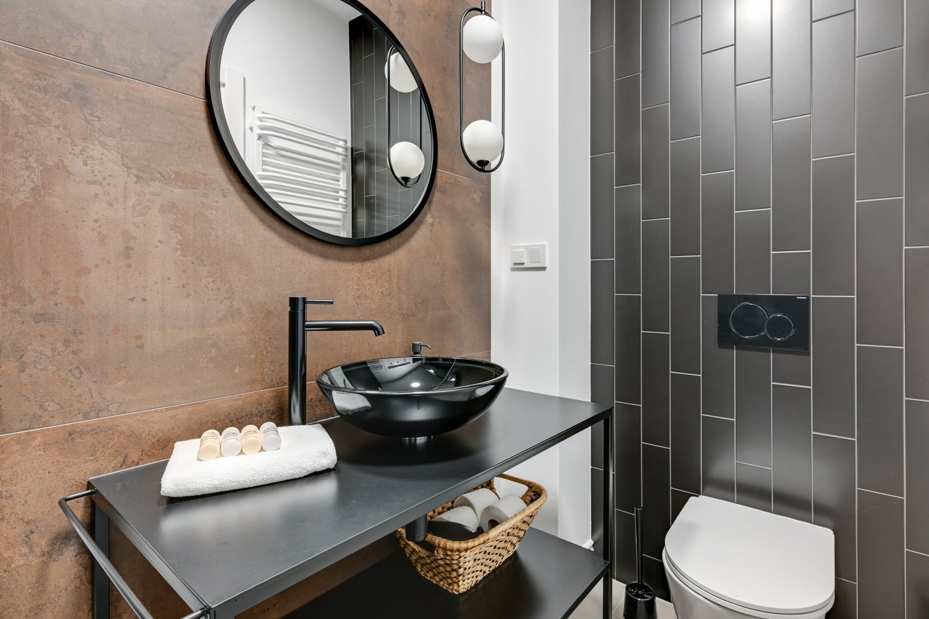 Nowoczesna łazienka z umywalką w kształcie misy, czarną armaturą i okrągłym lustrem, brązowe płytki w tle oraz szare płytki na ścianie z toaletą podwieszaną.