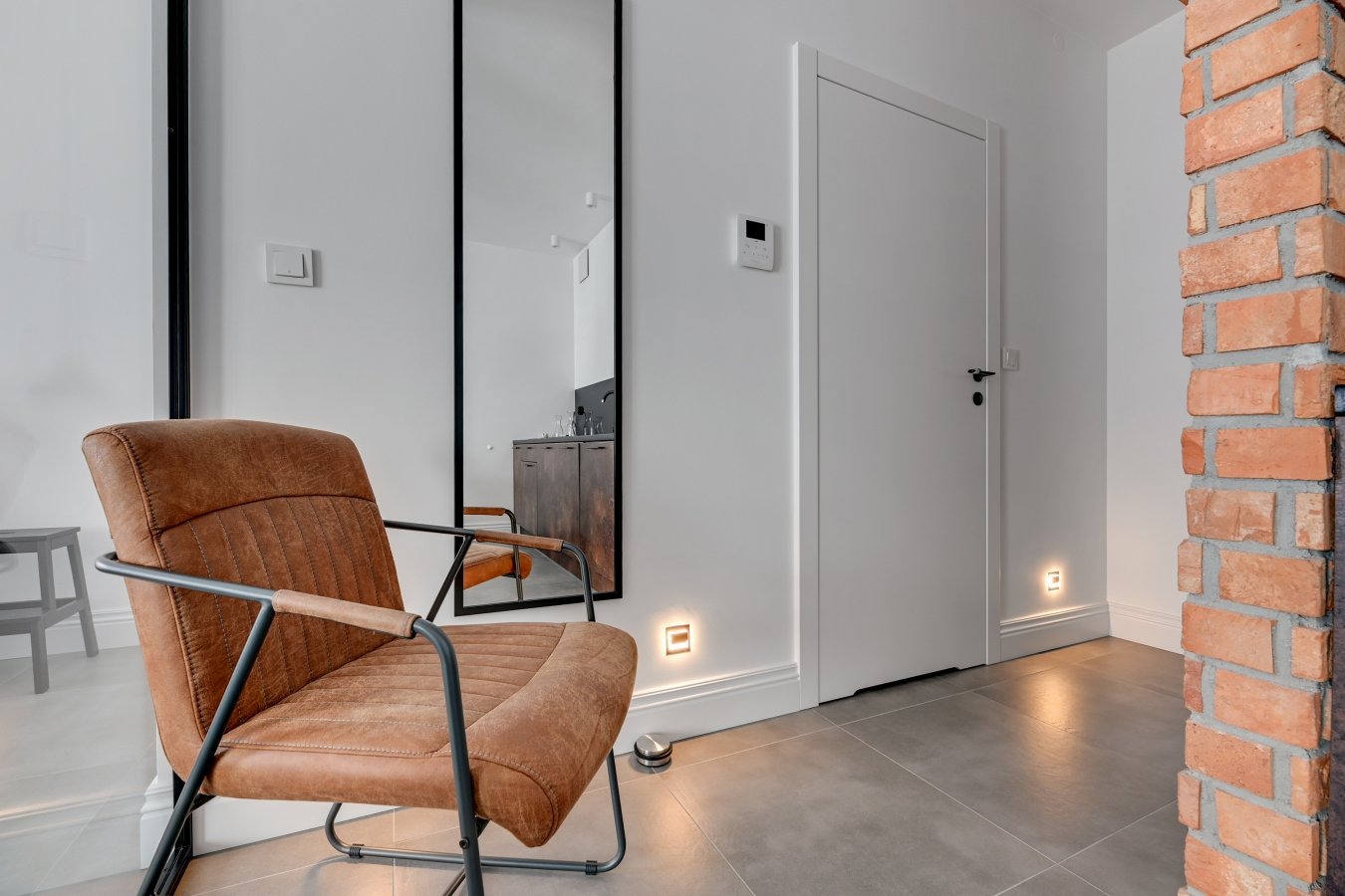 Wnętrze z fotelem w stylu vintage, lustrem w czarnej ramie, białą ścianą z ceglanym narożnikiem i podłogowymi lampkami.