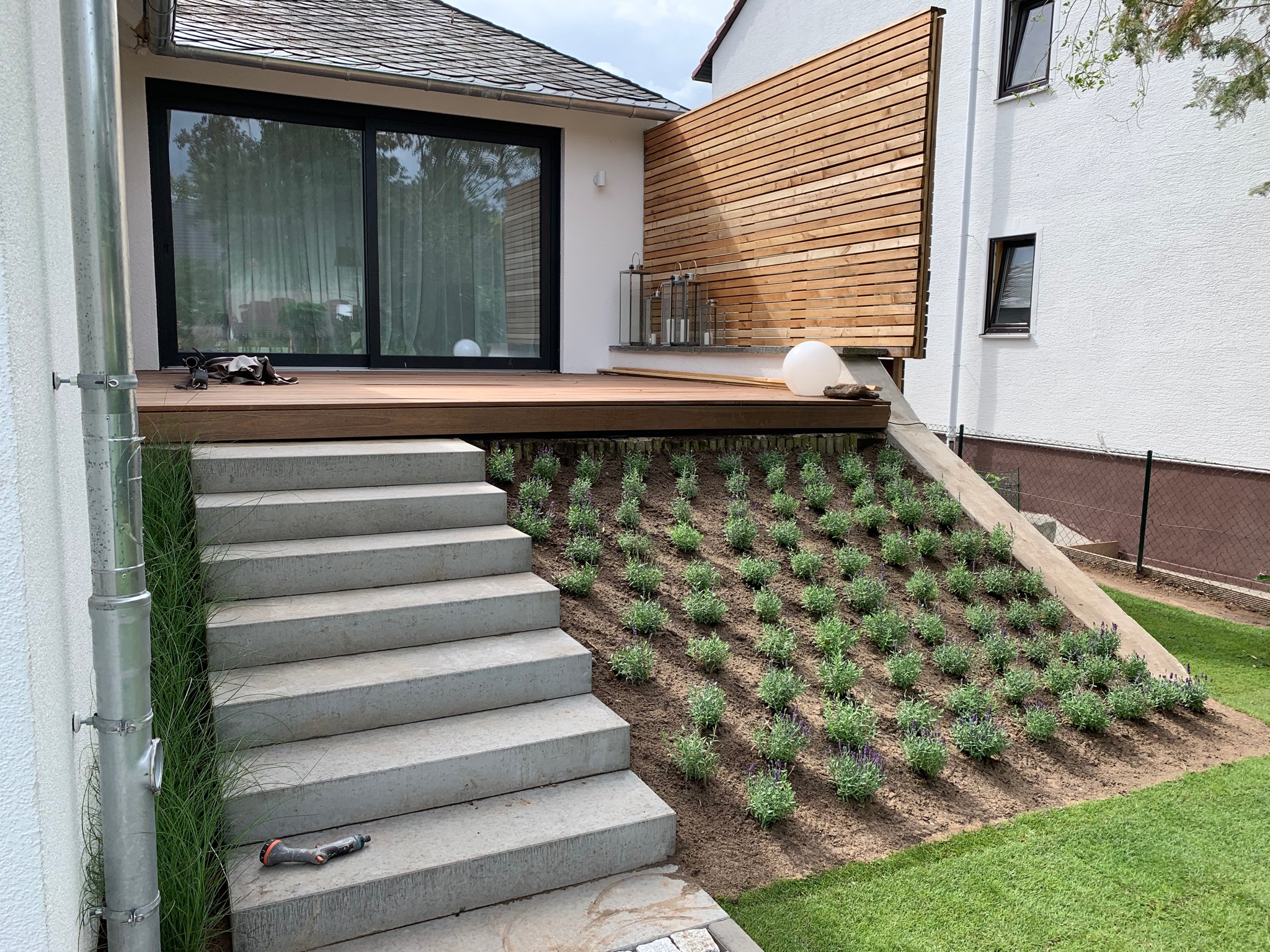 Nowoczesny taras z drewnianą podłogą i ażurową ścianą, schody z betonu prowadzą w dół, świeżo posadzona lawenda na skarpie, zraszacz leżący na schodach.