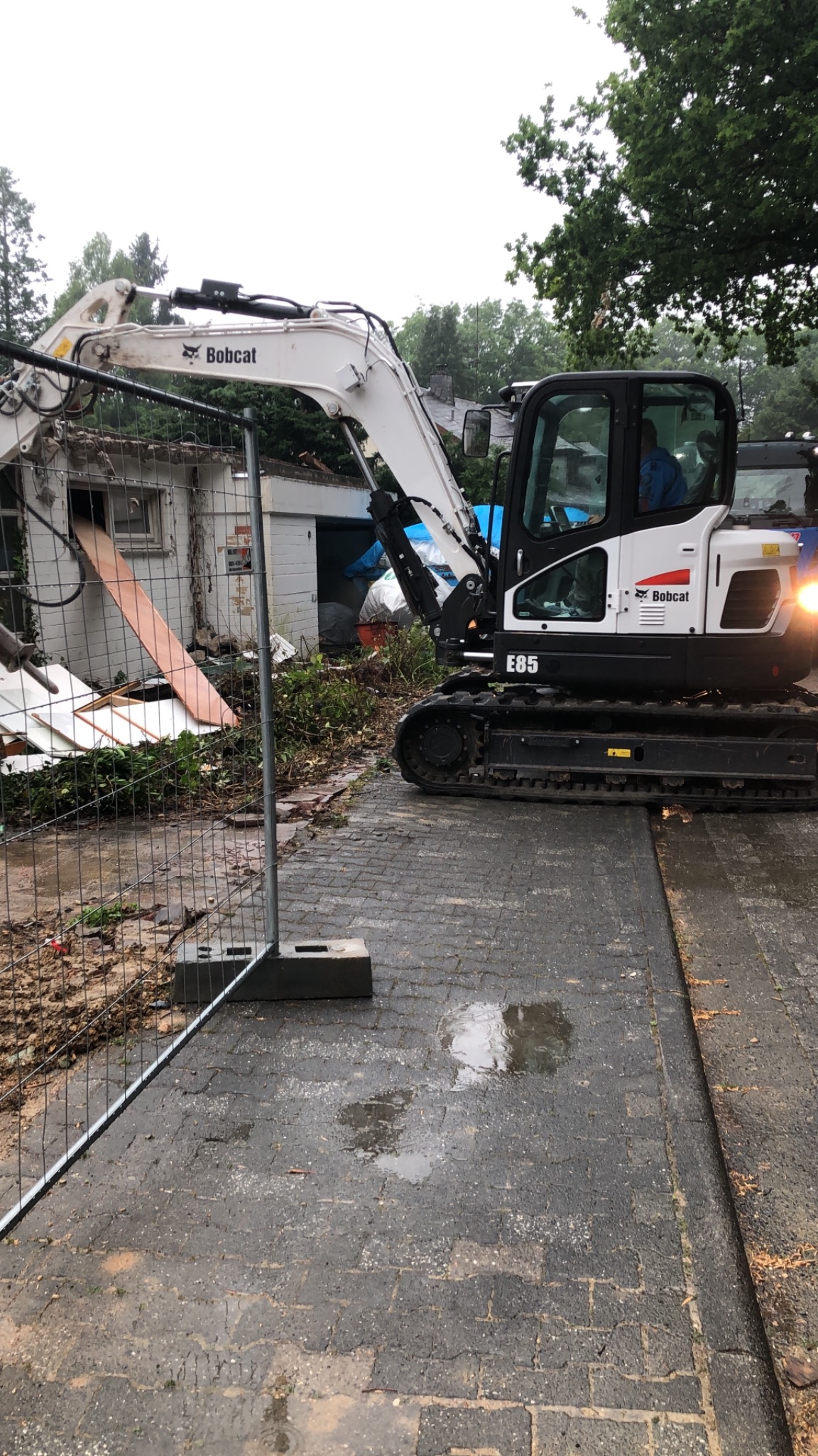 Biała minikoparka Bobcat E85 na mokrej, brukowej nawierzchni, w tle częściowo zburzony budynek z białej cegły i prowizoryczne ogrodzenie z siatki.