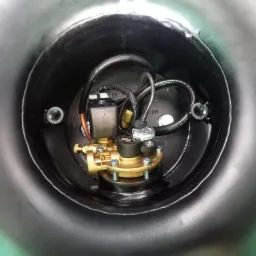 Zbliżenie na złoty reduktor ciśnienia gazu z przewodami i czarną obudową zbiornika w tle, widoczne śruby mocujące.