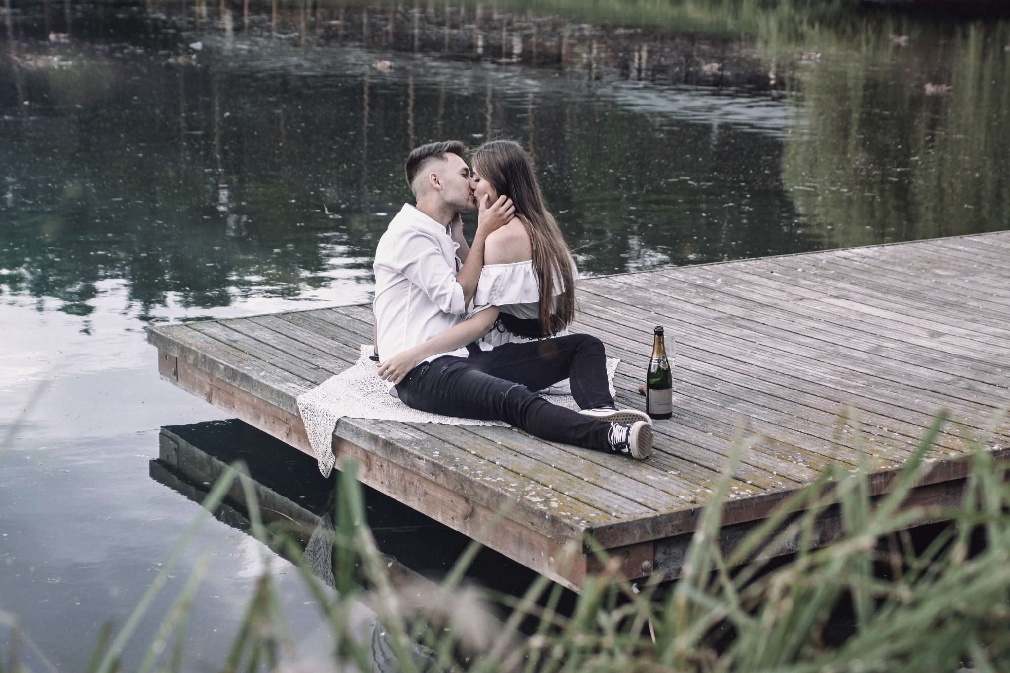 Zakochana para całuje się siedząc na drewnianym pomoście nad jeziorem, obok leży butelka szampana i kieliszek.