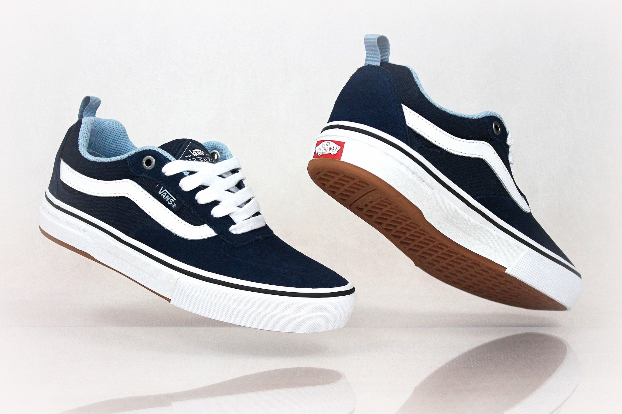 Dwa granatowe buty Vans z białymi paskami i sznurowadłami, unoszące się nad błyszczącą powierzchnią, ukazujące podeszwę z brązowego kauczuku.