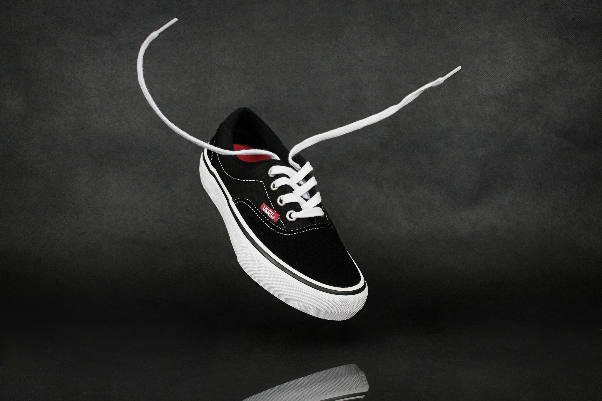 Lekko uniesiony czarny but Vans z białymi sznurówkami, z których jeden zwisa, na ciemnoszarym tle z subtelnym odbiciem pod spodem.