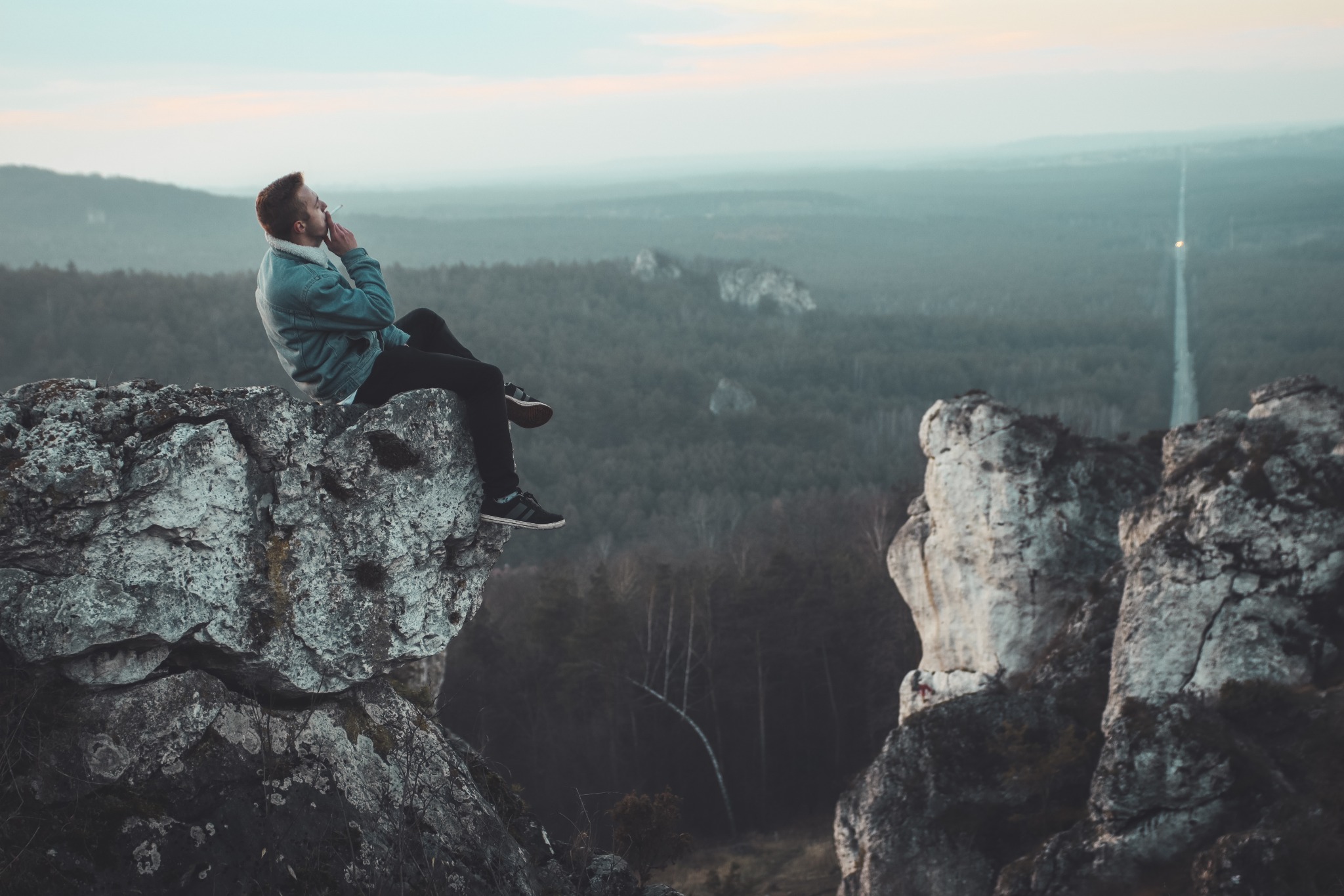 Mężczyzna w jeansowej kurtce siedzi na skalnym występie, paląc papierosa z widokiem na zalesione wzgórza i odległą drogę w porannym świetle.