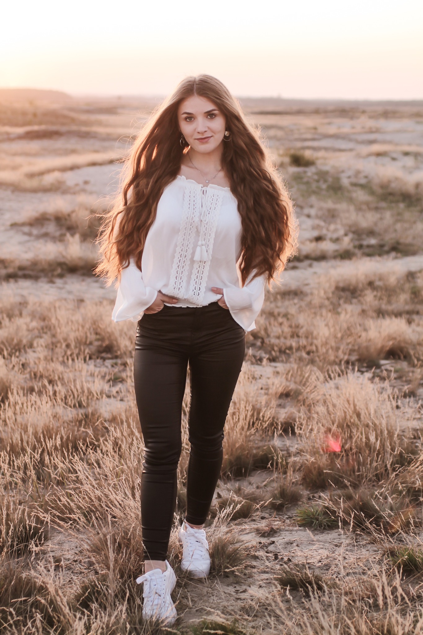 Portret kobiety w białej bluzce z długimi, falowanymi włosami na tle piaszczystego krajobrazu o zachodzie słońca.