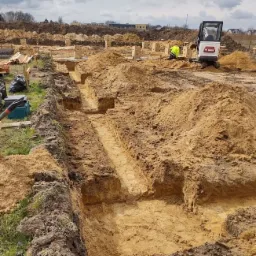 Głębokie wykopy pod fundamenty domu jednorodzinnego, widoczny mini-koparka Bobcat i pracownik w żółtej kamizelce, w tle kolejne wykopy i drewniane szalunki.