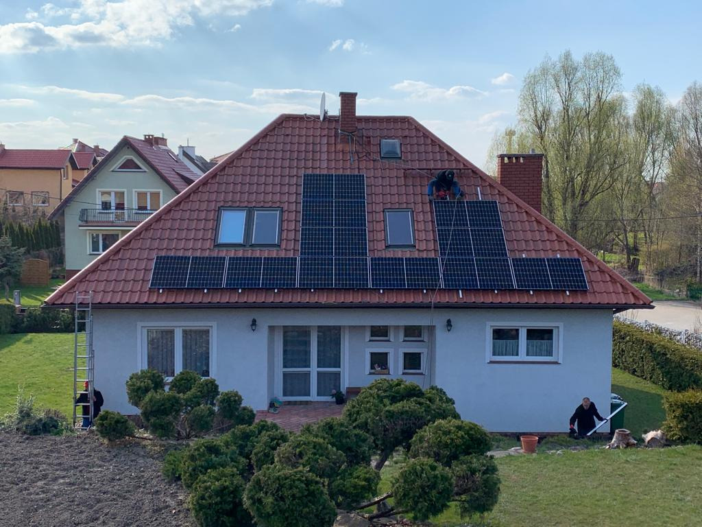 Montaż paneli słonecznych na dachu domu jednorodzinnego z czerwonej dachówki, widoczny pracownik w stroju roboczym na dachu oraz drabina oparta o ścianę, ogród z przodu domu.