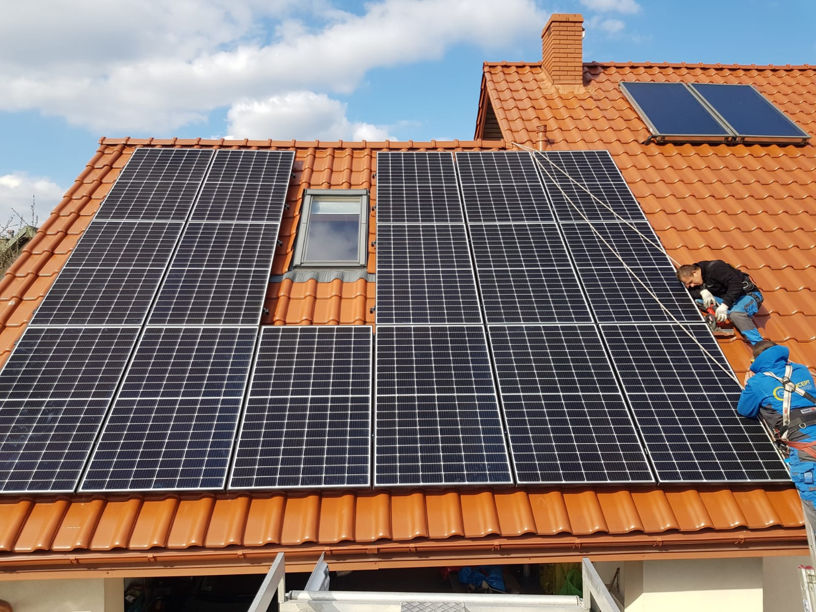 Montaż paneli słonecznych na dachu z czerwonej dachówki, widoczni dwaj monterzy w ubraniach roboczych i uprzężach, jeden z wkrętarką, okno dachowe i komin w tle.