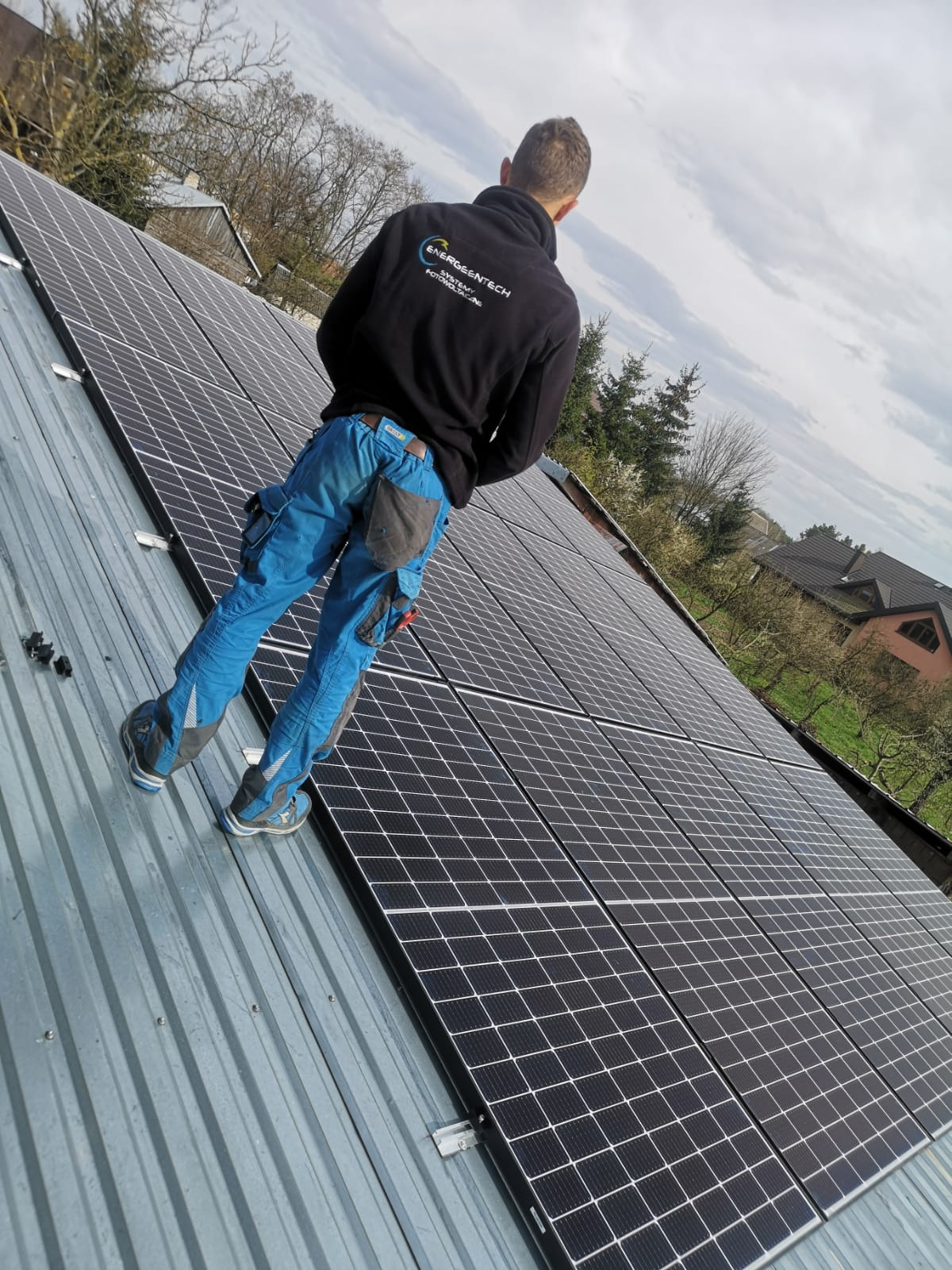 Pracownik w czarnej bluzie z logo firmy montuje panele słoneczne na blaszanym dachu, widok z dołu na pochmurne niebo.