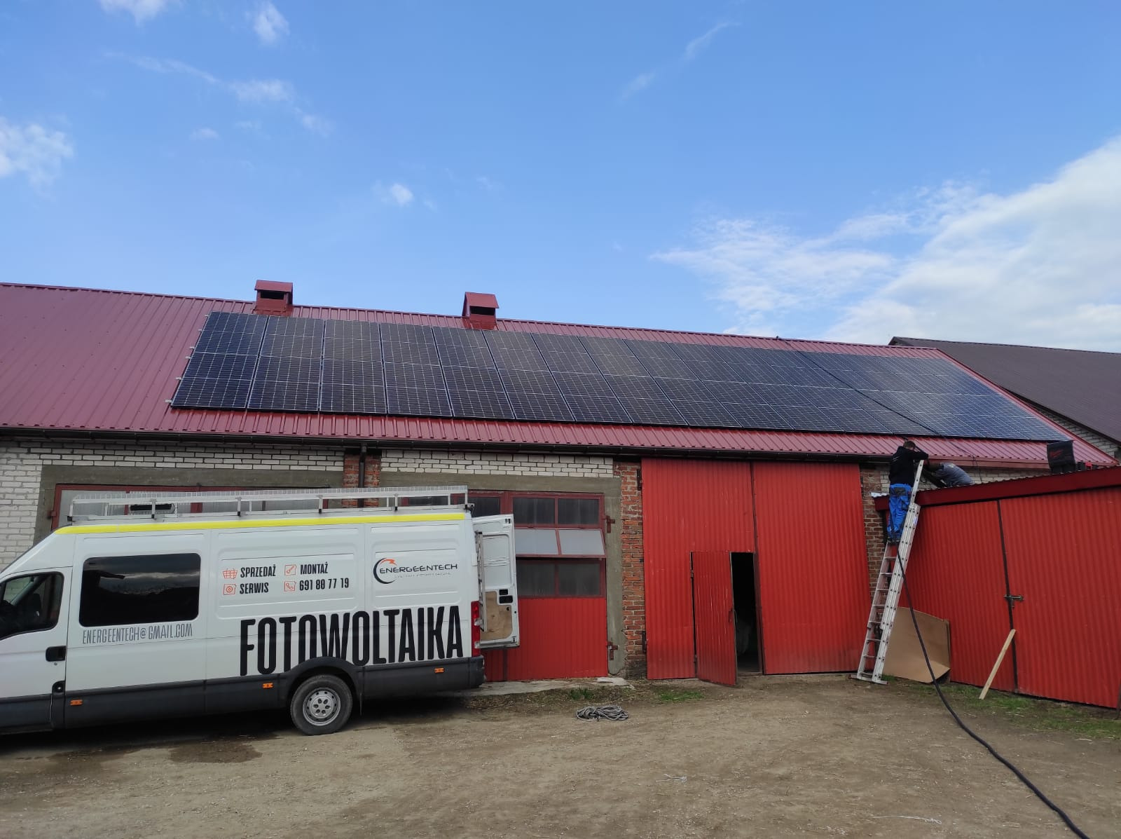Montaż paneli słonecznych na czerwonym, blaszanym dachu budynku gospodarczego; pracownik na drabinie, obok zaparkowany biały bus z napisem 'Fotowoltaika' i danymi kontaktowymi firmy.