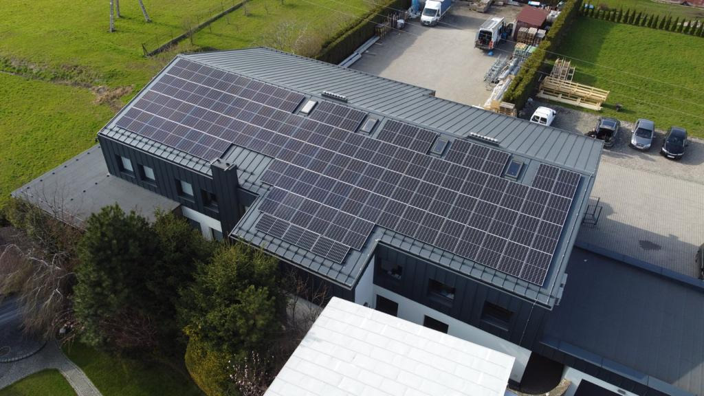 Widok z góry na budynek z zainstalowanymi panelami fotowoltaicznymi na szarym dachu, obok zaparkowane samochody i zielony trawnik.