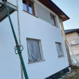 Świeżo otynkowana elewacja domu w Węgierskiej Górce, z oknami zabezpieczonymi folią, widoczny fragment rusztowania i niezagospodarowany teren wokół.